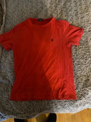 Röd t-shirt från Polo Ralph Lauren - Snygg röd t-shirt från Polo Ralph Lauren med klassisk svart logga broderad på bröstet. Modellen är kortärmad och har en rund halsringning. Perfekt för dig som gillar enkel och stilren design med en touch av street.