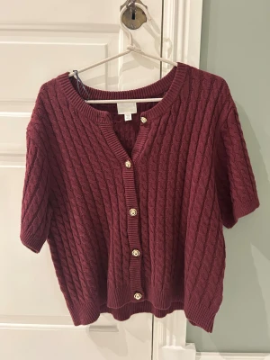 Vinröd kabelstickad kofta från H&M - Vinröd kabelstickad kofta från H&M med korta ärmar och snygga guldfärgade knappar framtill. Koftan har rund halsringning och ribbade muddar vid ärmslut och nederkant. Perfekt för lager-på-lager och en chill höstlook. Aldrig använd och vem som helst kan ha beroende på hur man vill att den ska sitta