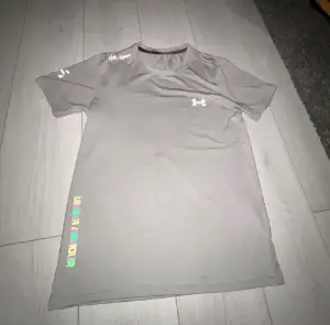 Snygg grå t-shirt från Under Armour med tryck på bröstet och ärmen. Modellen har rund hals och korta ärmar, perfekt för gymmet eller löprundan. Materialet är lätt polyester som andas och transporterar bort fukt. Diskret logga och stilren design.
