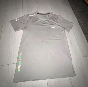 Grå Under Armour t-shirt för träning - Snygg grå t-shirt från Under Armour med tryck på bröstet och ärmen. Modellen har rund hals och korta ärmar, perfekt för gymmet eller löprundan. Materialet är lätt polyester som andas och transporterar bort fukt. Diskret logga och stilren design.