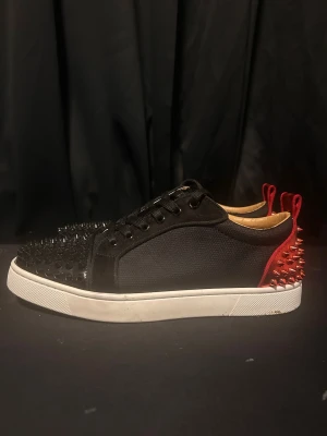 Louboutins  - Unika svarta sneakers med grova svarta nitar på tån och röda metalliska nitar på hälen. Skorna har snörning, vit platt sula och en cool kontrast mellan svart textil och röd detalj bak. Perfekt för dig som vill sticka ut med edgy stil. Louboutins stolek 40,41 bra skick alla spikar finns 