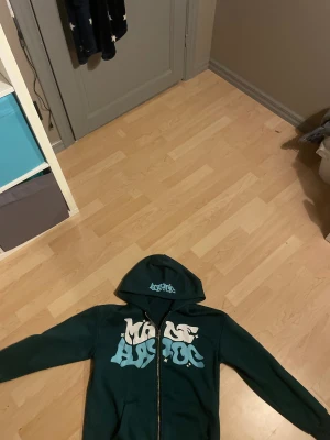 Grön hoodie med tryck från Magic - Mörkgrön hoodie med stor vit och ljusblå grafisk text på bröstet och mindre tryck på huvan. Tröjan har dragkedja framtill, huva och långa ärmar. Materialet är mjuk polyester med lite stretch för extra komfort. Perfekt för dig som gillar streetwear vibes.