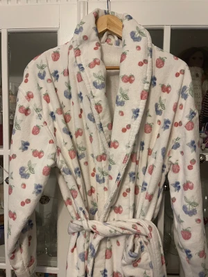 Fruktmönstrad frottérock från Djerf Avenue - Säljer en vit frottérock från Djerf Avenue med gulligt fruktmönster i rött, blått och grönt. Rocken har lång ärm, bred krage och knyts med bälte i midjan. Perfekt för chill hemma och har en riktigt cozy vibe.