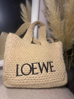 Beige handväska från Loewe - Säljer en beige handväska från Loewe med grovstickad struktur och tydlig svart logotyp på framsidan. Väskan har två rejäla handtag och är tillverkad i stickat material som ger en chill och naturlig vibe. Perfekt storlek för att få plats med allt du behöver.