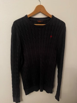 Grå kabelstickad tröja Morris M - Svart kabelstickad tröja från Morris i storlek M. Tröjan har ett diskret rött broderat emblem på bröstet och rund hals. Stickad struktur med klassiskt mönster och långa ärmar. Perfekt för dig som gillar stilrena och tidlösa plagg. M och S