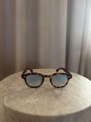 Noa 02 - Bilderna visar ett par solglasögon med sköldpaddsmönstrad båge och blåtonade glas. Ingen väst syns på bilderna.