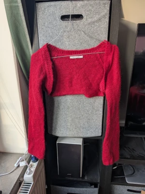 Röd croppad stickad bolero Message - Croppad röd stickad boleri från Message med lång ärm och bred halsringning. Tröjan är tillverkad i ett mohair stickat material som känns fluffigt och skönt.