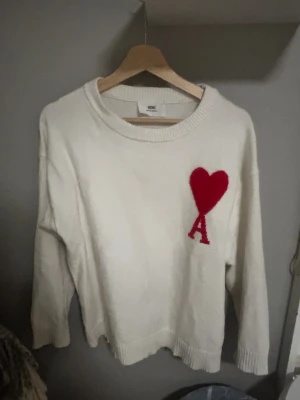 Vit stickad tröja från Ami - Vit stickad tröja från Ami Alexandre Mattiussi med rund hals och ribbade muddar. På bröstet finns ett stort rött hjärta och bokstaven A som ger en unik och trendig look. Perfekt för dig som gillar stilrena detaljer och vill sticka ut. Om du behöver något annat fråga bara. 