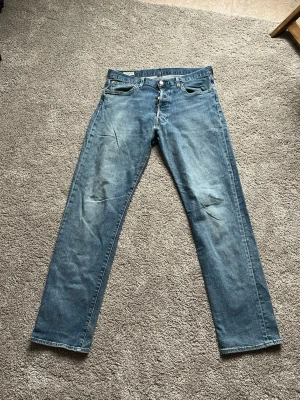 Levi's 501 blå jeans, straight fit - Klassiska Levi's 501 jeans i ljusblå tvätt med raka ben och fem fickor. Jeansen har knappgylf och den ikoniska läderpatchen bak i midjan. Materialet är slitstark bomull och passformen är straight. Perfekt för dig som gillar tidlös denimstil.