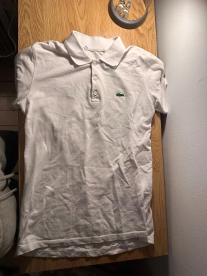Vit pikéskjorta från Lacoste - Säljer en klassisk vit pikéskjorta från Lacoste med den ikoniska gröna krokodilen broderad på bröstet. Skjortan har krage, tre vita knappar framtill och är gjord i mjuk bomull. Perfekt för dig som gillar stilren och enkel design.