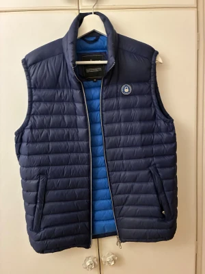 Mörkblå dunväst från Sail Racing - Säljer en mörkblå dunväst från Sail Racing i storlek M. Västen är i bra skick och knappt använd, ganska tunt material så passar perfekt att ha till vår/sommar, pris kan diskuteras!!