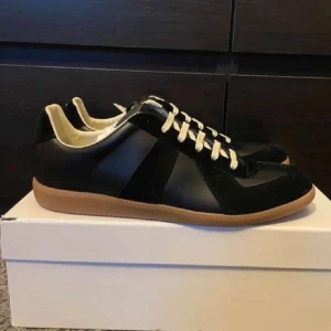 Svarta maison margiela gats skor - Säljer ett par svarta maison margiela (helt nya aldrig använda) sneakers med beige snörning och gummisula i brunt. Skorna har en snygg mix av skinn och mocka på ovandelen, rund tå och platt sula. Insidan är ljus och mjuk för extra komfort. Perfekt för dig som gillar stilrena och klassiska sneakers. Storleken är 44 men passar mindre som 42-43