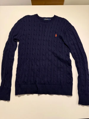 Marinblå stickad tröja Polo Ralph Lauren - OBS: tröjan är omgjord för att passa storlek M, därmed inte helt naturlig passform men det märks inte. Säljer en marinblå kabelstickad tröja från Polo Ralph Lauren i storlek XL. Tröjan har rund hals, långa ärmar och ett orange broderat logomärke på bröstet. Ribbstickade muddar vid ärmslut och nederkant. Perfekt för dig som gillar klassisk stil.