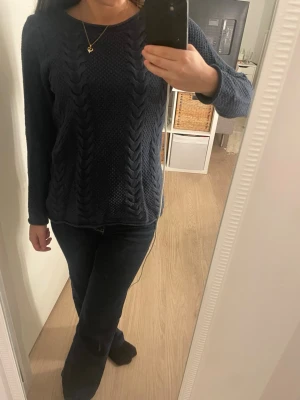 Mörkblå stickad tröja med kabelmönster - Säljer en mörkblå stickad tröja med snyggt kabelmönster framtill. Tröjan har rund hals och långa ärmar, perfekt för kyliga dagar. Materialet är mjukt och ger en skön känsla mot huden. Passar dig som gillar klassisk och enkel stil.