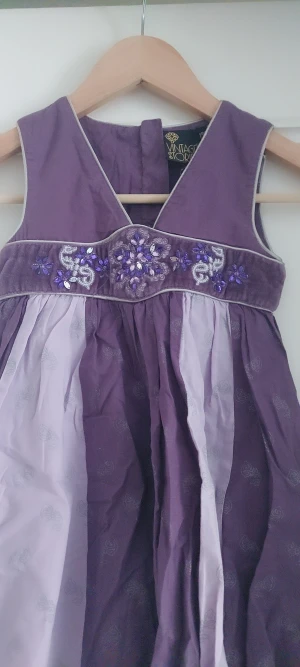 Lila kortklänning från Kappahi, barnkläning storlek 122 - Supersöt lila kortklänning från Vintage Stories med bred midjedel dekorerad med pärlor och broderade blommor. Klänningen har v-ringad hals och är ärmlös, med skira paneler i ljuslila och lila tyg. Perfekt för dig som gillar detaljer och färg!