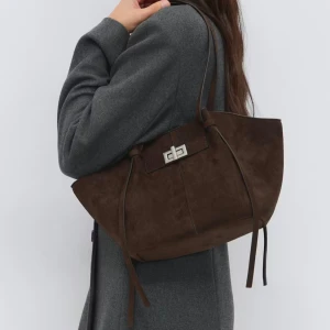 Brun mocka axelväska med snörning - Snygg brun axelväska i mocka med långa axelband och dekorativa snörningsdetaljer på sidorna. Väskan har en cool metallspänne framtill och en mjuk, formad siluett som ger en trendig vibe. Perfekt storlek för att få plats med allt du behöver. Midi long strap bag from gina tricot