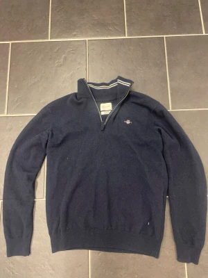 Mörkblå GANT half zip tröja i ull - Snygg mörkblå tröja från GANT i superfin lammull, med halv dragkedja och ribbad krage med vita ränder. Klassisk broderad GANT-logga på bröstet. Perfekt för dig som gillar stilrena och bekväma plagg med premiumkänsla.