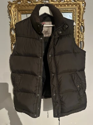 Svart dunväst från Esprit - Säljer en svart dunväst från Esprit med hög krage och breda quiltade paneler. Västen har både dragkedja och tryckknappar framtill samt två fickor med lock och knapp. Perfekt för lager-på-lager och snygg till streetwear.