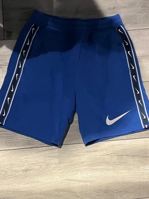 Blåa Nike shorts  - Säljer ett par blå Nike shorts med vita loggor längs sidorna och en stor vit swoosh framtill. Inga defekter eller tecken på användning. De har elastisk midja och är gjorda i mjuk bomull. Snygg design med kontrastband på båda benen. Pris kan diskuteras vid snabb affär. 
