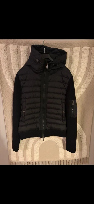 Moncler cardigan  - Säljer en svart pufferjacka från Moncler med stickade ärmar och detaljerade dragkedjor. Jackan har hög krage, huva och Moncler-logga på ena ärmen. Perfekt för kyliga dagar och har en snygg, modern look. Liten fläck på insidan men syns inte vid användning 