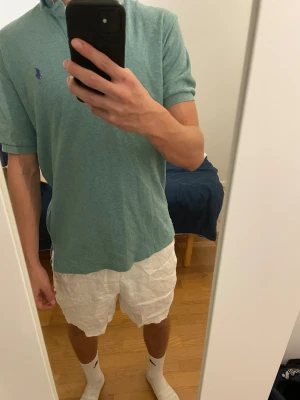 Ralph Lauren piké - Snygg grön pikétröja från Polo Ralph Lauren med klassisk krage och två knappar framtill. Tröjan har korta ärmar och den ikoniska lila Polo-loggan broderad på bröstet. Perfekt för dig som gillar stilrena och bekväma plagg.