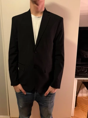 Svart kavaj från H&M Slim Fit - Säljer en svart kavaj från H&M i slim fit-modell. Kavajen har klassisk krage, två knappar framtill och fickor med lock. Perfekt för dig som vill ha en stilren och enkel look. Materialet är mjukt och följsamt, vilket gör den bekväm att bära.