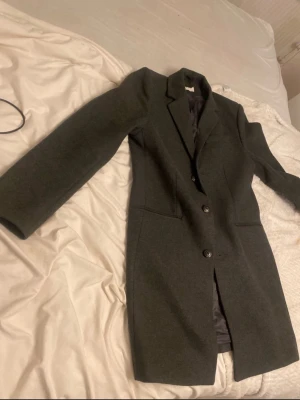 Mörkgrå kappa med knappar - Snygg mörkgrå kappa med klassisk krage och två knappar framtill. Jackan har raka, långa ärmar och två diskreta fickor på sidorna. Perfekt för dig som gillar stilrena och tidlösa plagg. Materialet känns mjukt och har en ullig look. Original pris är runt 2 tusen från zalando