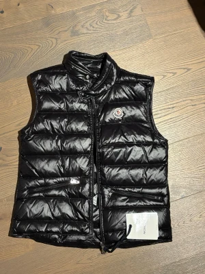 Svart dunväst från Moncler - Säljer en stilren och klassisk svart dunväst från Moncler. Västen är lätt, varm och perfekt att bära över hoodie eller tröja under höst/vår – eller som mellanlager på vintern. Aldrig avkänd 