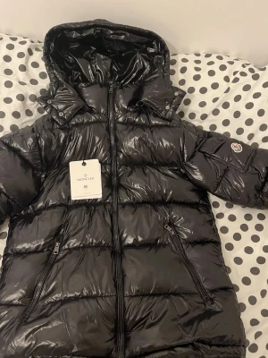 Svart Moncler pufferjacka med huva - Säljer en svart pufferjacka från Moncler med glansig finish och huva. Jackan har dragkedja framtill, två sidofickor med dragkedja och Moncler-logga på ärmen. Insidan har en cool serietidningspatch. Perfekt för kalla dagar och riktigt snygg streetstil.