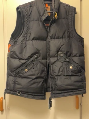 Svart dunväst från Parajumpers XL - Säljer en svart dunväst från Parajumpers i storlek XL. Västen har två snedställda fickor med dragkedja och knapp, hög krage med tryckknappar och detaljer i orange. Perfekt för kalla dagar och har en quiltad design som ger extra värme.