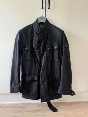 Belstaff Roadmaster  - Svart Roadmaster från Belstaff med klassisk krage, bälte vid halsen och flera fickor med tryckknappar. Materialet är polyester och bomull, perfekt för höst och vinter. Jackan är i ett mycket bra skick