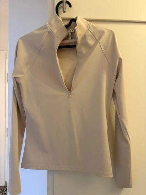 Beige långärmad polotopp från NLY Trend - Säljer en stilren beige polotopp från NLY Trend i storlek M. Toppen har lång ärm, hög krage och en snygg dragkedja framtill som kan justeras för att skapa en djupare v-ringning. Materialet är mjukt och stretchigt, perfekt för att matcha med jeans eller kjol.