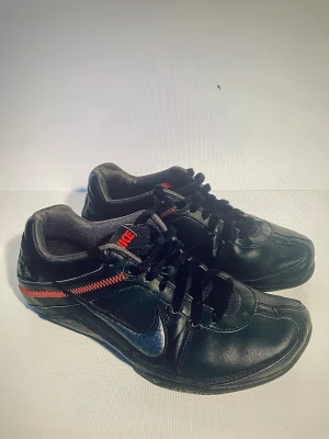 Svarta Nike sneakers Zoom Porto Leader - Nike sneakers i svart skinn med lågt skaft och rund tå. Snyggt röd zigzag-söm längs sidan och röd Nike-logga på plösen. Svarta skosnören och platt sula. Perfekt för dig som gillar stilrena och sportiga skor med en twist. Sparsamt använda. Storlek 42, innermått 26,5 cm.