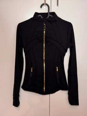 Svart träningsjacka från Lululemon NY - NY Snygg svart träningsjacka från Lululemon med guldiga dragkedjor och figurnära passform. Jackan har högt krage, långa ärmar och två fickor med dragkedja framtill. Köpt utomlands därav priset :-) 