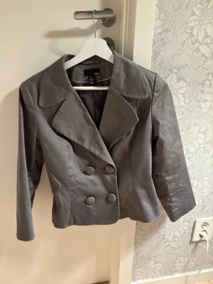 Grå kavajjacka från H&M, strl 36 - Snygg grå kavajjacka från H&M i storlek 36. Jackan har bred krage, dubbelknäppning med stora tygklädda knappar och långärmad design. Perfekt för dig som gillar stilrena och klassiska plagg med modern touch.