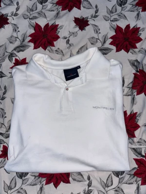 Vit polotopp från Montpelier XL - Säljer en klassisk vit polotopp från Montpelier i storlek XL. Toppen har krage, kort knappslå och diskret logga broderad på bröstet. Materialet är mjuk bomull som känns skönt mot huden. Perfekt för dig som gillar stilrena och enkla plagg.