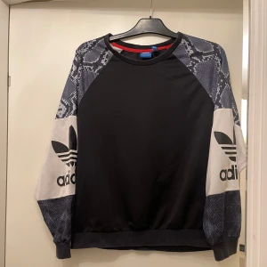Adidas sweatshirt med ormmönster - Svart Adidas sweatshirt med grå och vita detaljer på ärmarna, stora logotryck och coolt ormmönster. Tröjan har rund hals, ribbade muddar och lång ärm. Perfekt för dig som gillar streetwear och sportig stil.