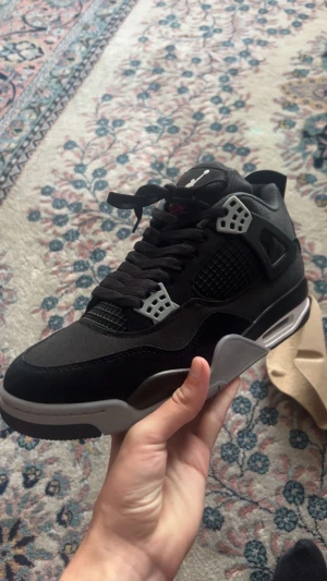 Nike Air Jordan 4 svart/grå sneaker - Nike Air Jordan 4 sneakers i svart och grått med röda detaljer på yttersulan. Skon har meshpaneler, snörning och Jumpman-logga på hälen och sulan. Materialet är en mix av syntet och mocka, med en platt sula och rund tå. Perfekt för dig som gillar klassiska och sportiga sneakers.