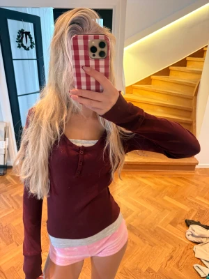 Vinröd långärmad topp med knappar - Säljer en vinröd långärmad topp med rundad ringning och tre knappar framtill. Modellen är figurnära och toppen är i ett mjukt, stretchigt material som känns skönt mot huden. Perfekt att styla med jeans eller kjol för en enkel look.