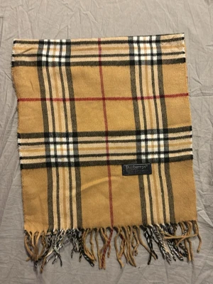 Rutig Burberrys halsduk i lammull - Säljer en klassisk rutig halsduk från Burberrys i beige, svart, vit och röd. Halsduken är tillverkad i 100% lammull och har fransar i ändarna. Den ikoniska rutiga designen ger en tidlös vibe och passar perfekt till höst och vinter. Inga defekter. Hör av dig vi frågor eller funderingar🤩😃👍.