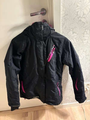 Svart vindjacka från Pyelix med rosa detaljer - Svart vindjacka från Pyelix med snygga rosa dragkedjor och detaljer. Jackan har huva, flera fickor med dragkedja och är perfekt för blåsiga dagar. Materialet är slitstark polyester och jackan har en sportig look som sticker ut. Den kan även passa som skidjacka. 