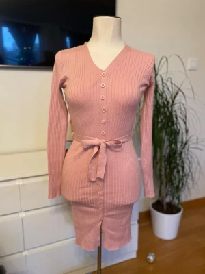 Rosa ribbad stickadklänning med knyt - Säljer en söt rosa ribbad klänning med v-ringning, långa ärmar och knappar hela vägen framtill. Klänningen har ett matchande bälte i midjan som knyts och ger en snygg siluett. Perfekt för dig som gillar stilrena detaljer och mjukt material.