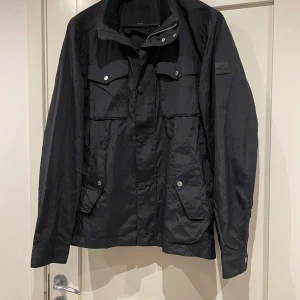 Svart vindjacka från J.Lindeberg  - Svart vindjacka från J.Lindeberg med flera fickor framtill, knappar och dragkedja. Jackan har en stilren design med patch på ärmen och detaljer i silver. Perfekt för dig som gillar funktion och snygg stil. Storlek xl men sitter mer som L