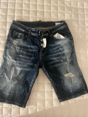 Dsquared2 jeansshorts med slitningar - Säljer ett par mörkblå jeansshorts från Dsquared2 med coola slitningar, färgstänk och knappar framtill. Klassisk femficksdesign och en grön Dsquared2-label bak. Perfekta för dig som gillar streetstyle och vill sticka ut lite extra.