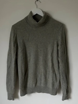 Grå polotröja i ull från Uniqlo - Säljer en stilren grå polotröja från Uniqlo i 100% ull. Tröjan har en hög, ribbad krage och långa ärmar med ribbade muddar. Perfekt för dig som gillar klassisk och enkel design med mjukt material som håller värmen. 5L16