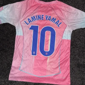  Lamine Yamal tröja  - Säljer en FC Barcelona fotbollströja med Lamine Yamal och nummer 10 på ryggen. Tröjan är rosa med ljusblå detaljer.