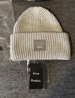 Grå stickad mössa från Acne Studios - Säljer en snygg grå mössa från Acne Studios i 100% ull. Mössan är ribbstickad med uppvikt kant och har Acne Studios klassiska ansiktslogga framtill. Perfekt för dig som vill ha en clean och stilren look under kyliga dagar.