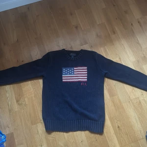Mörkblå stickad tröja Polo Ralph Lauren - Säljer en mörkblå stickad tröja från Polo Ralph Lauren med amerikansk flagga och RL-broderi på bröstet. Tröjan har rund hals och långa ärmar med ribbade muddar vid ärmslut och nederkant. Perfekt för dig som gillar klassisk stil.