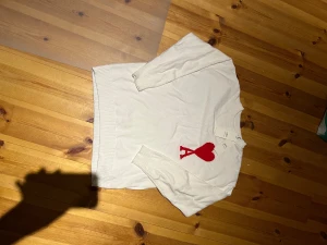 Vit stickad tröja från Ami paris - Vit stickad tröja från Ami Alexandre Mattiussi med röd hjärt- och A-detalj på bröstet. Tröjan har rund hals, ribbade muddar och är långärmad. Tillverkad i Portugal av mjuk bomull, perfekt för dig som gillar stilrena och coola detaljer.