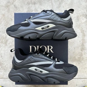 Svarta chunky sneakers från Dior - Säljer ett par svarta Dior B22 med cool design och detaljerad sula. Skorna har meshpaneler, skinn och syntetmaterial, samt snörning och rund tå. Perfekta för dig som gillar exklusiva skor. Dessa är i storlek 43. Använda men i mycket bra skick. 